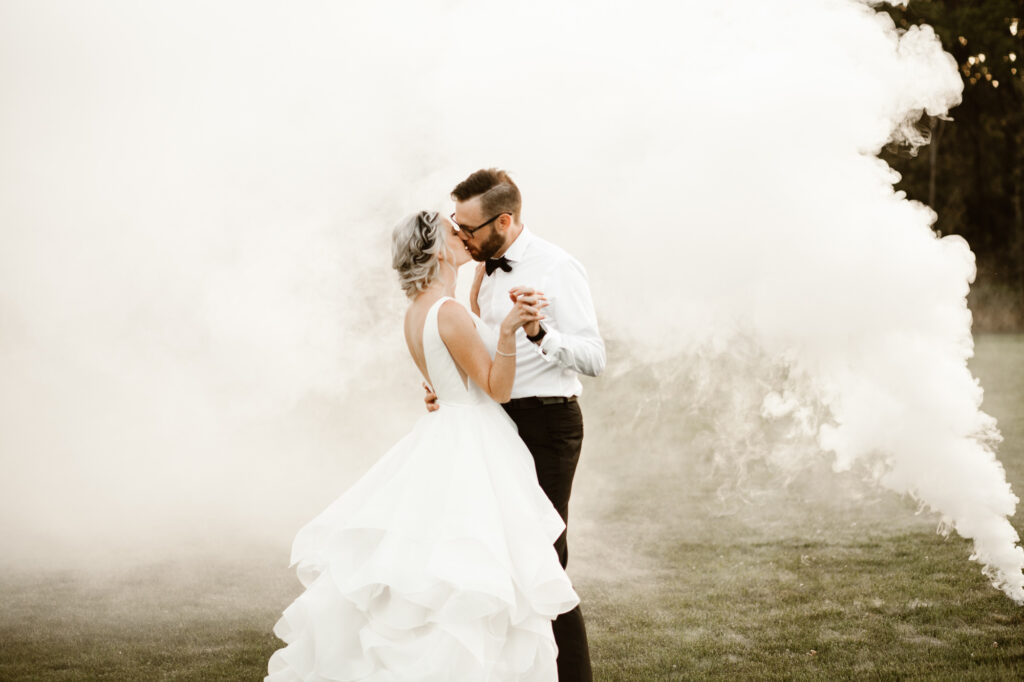 Married couple kissing against smoke bomb backdrop at Maple Meadows wedding venue.
