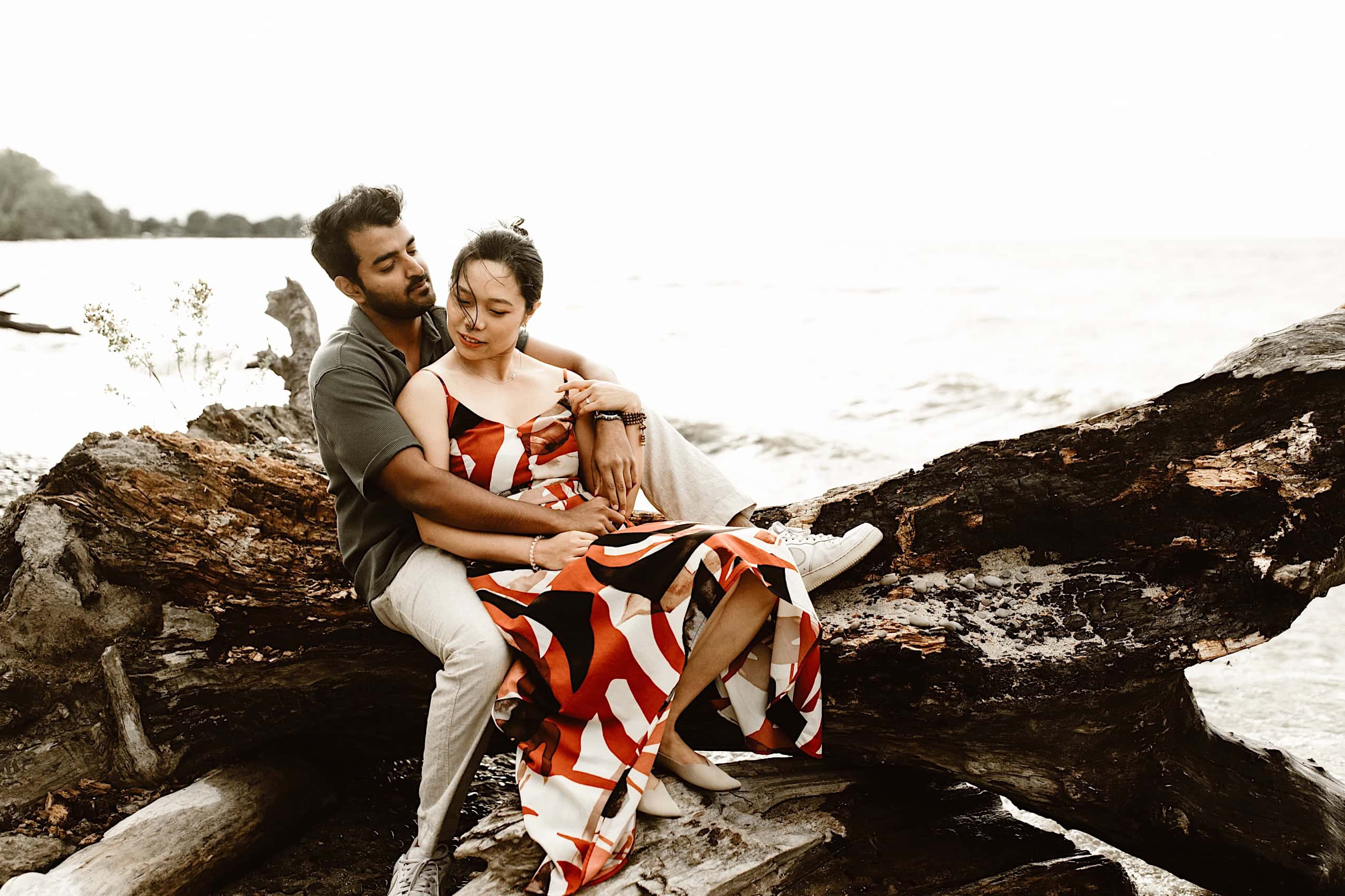 engaged couple holding each other on a piece of driftwwod by lake ontario